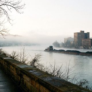 Steam rising off a wide gray river at dawn beside a bare paved walkway and a low stone wall