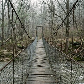 A narrow wooden footbridge hung on cables above a fast brown creek between bare early spring trees
